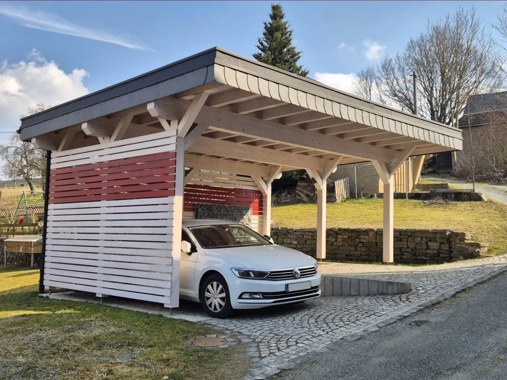 Moderner Holzcarport mit Flachdach und seitlicher Lamellenverkleidung in Weiß und Rot, darunter ein weißer Pkw auf gepflasterter Zufahrt vor ländlichem Grundstück.