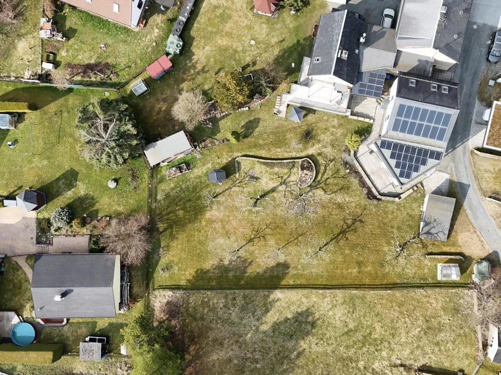 Drohnenaufnahme eines großen Grundstücks mit Einfamilienhaus, Garten, Obstbäumen und Nebengebäuden, inklusive modernem Carport mit Photovoltaikdach in ländlicher Umgebung.