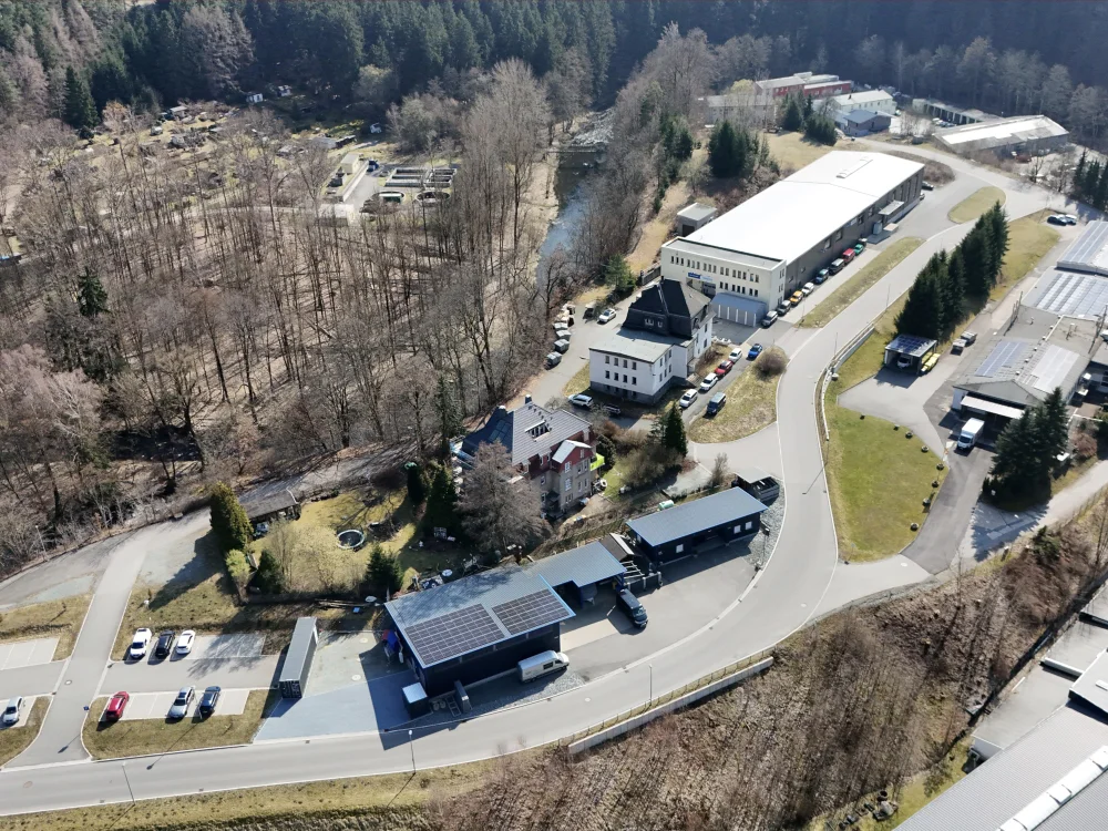 Drohnenaufnahme eines Gewerbegrundstücks mit moderner Holzhalle, Carport mit Photovoltaikanlage und umliegender Bebauung in ländlicher Umgebung.