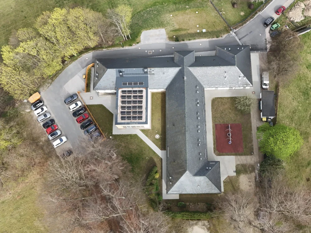 Drohnenaufnahme eines Gebäudekomplexes mit Parkplatz, Außenanlagen und Photovoltaikanlage auf dem Dach in ruhiger, grüner Umgebung.