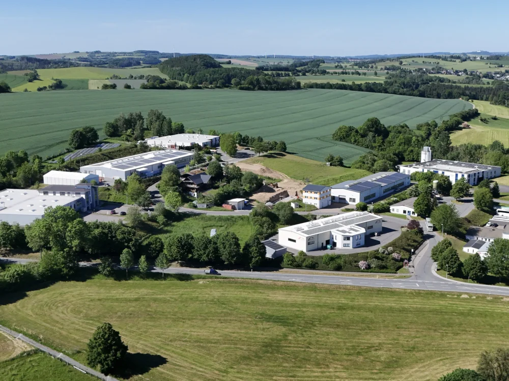 Drohnenaufnahme eines Gewerbegebiets mit modernen Produktionshallen, Photovoltaikanlagen auf den Dächern und umliegender ländlicher Landschaft.