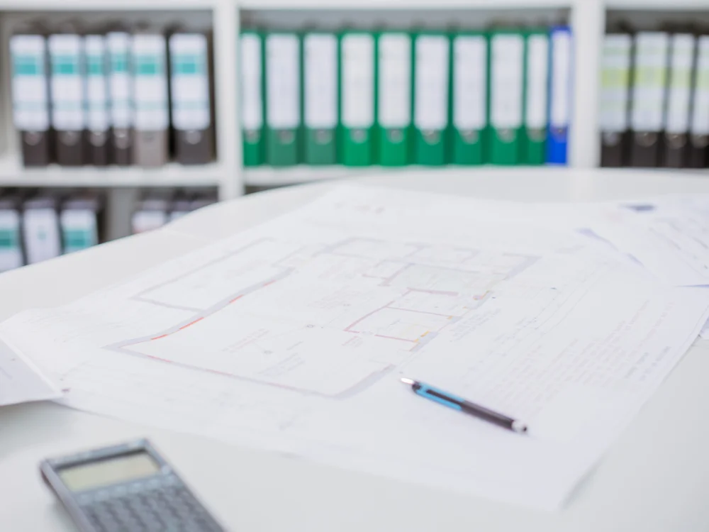 Bauplan und technische Zeichnungen auf Schreibtisch mit Taschenrechner und Ordnern im Hintergrund, Darstellung von Bauplanung und Projektvorbereitung im Ingenieurbüro.