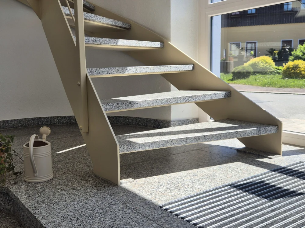 Moderne Innentreppe mit Natursteinstufen und Stahlkonstruktion im hochwertigen Innenausbau, kombiniert mit großen Fensterflächen und hellem Eingangsbereich.