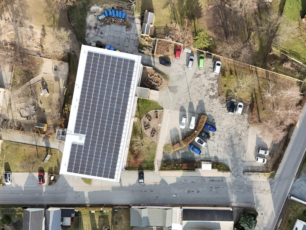 Drohnenaufnahme eines Gebäudekomplexes mit großflächiger Photovoltaikanlage auf dem Dach, Parkplatz und Außenanlagen in ländlicher Umgebung.