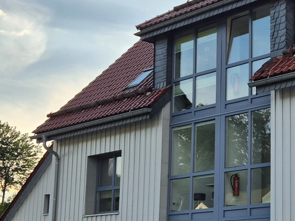 Moderne Hausfassade mit Holzverkleidung, großen Fensterflächen und Steildach mit Ziegeldeckung, kombiniert mit Gaube und Dachfenster im hochwertigen Wohnbau.