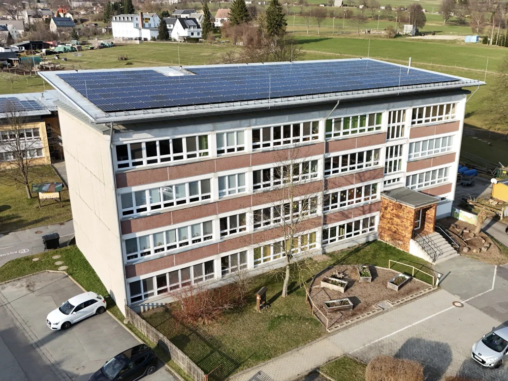 Drohnenaufnahme eines Schul- oder Verwaltungsgebäudes mit großflächiger Photovoltaikanlage auf dem Flachdach, Außenanlagen und Parkflächen in ländlicher Umgebung.