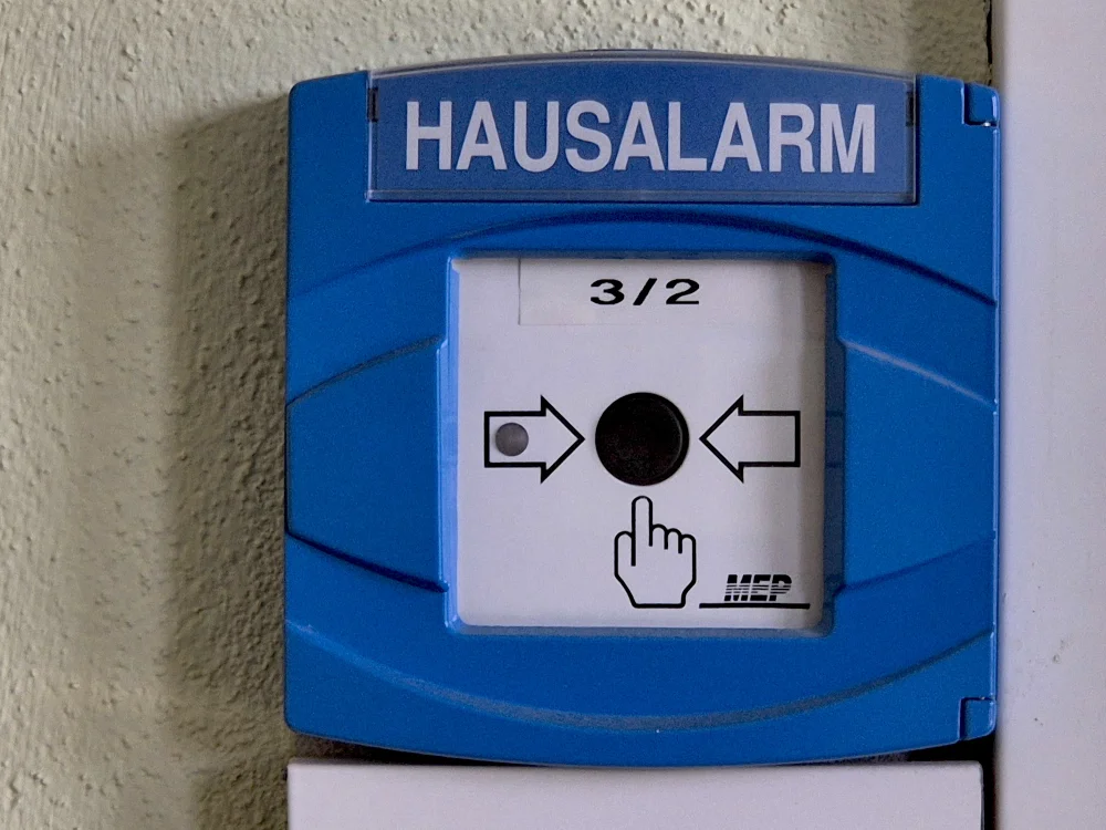 Hausalarm-Brandmelder mit Druckknopf zur manuellen Auslösung im Innenbereich, Teil moderner Brandmeldeanlage für schnelle Alarmierung im Notfall.