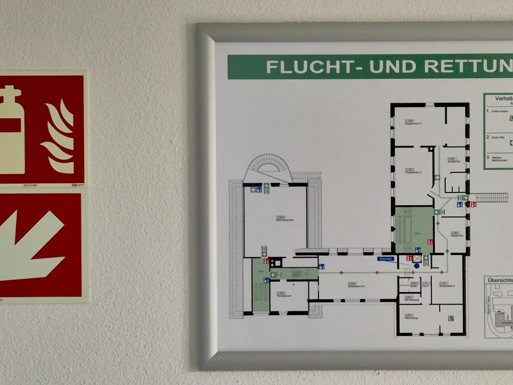 Flucht- und Rettungsplan mit Grundrissdarstellung und Sicherheitskennzeichnungen im Innenbereich, ergänzt durch Brandschutzzeichen für Feuerlöscher und Notausgänge.