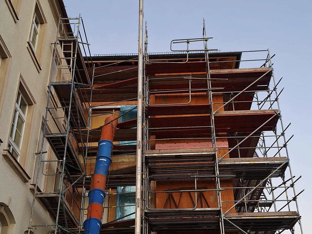 Baugerüst an mehrstöckigem Gebäude mit Mauerwerksarbeiten und Materialrutsche, eingesetzt für Fassadenbau und Rohbauarbeiten im Hochbau.
