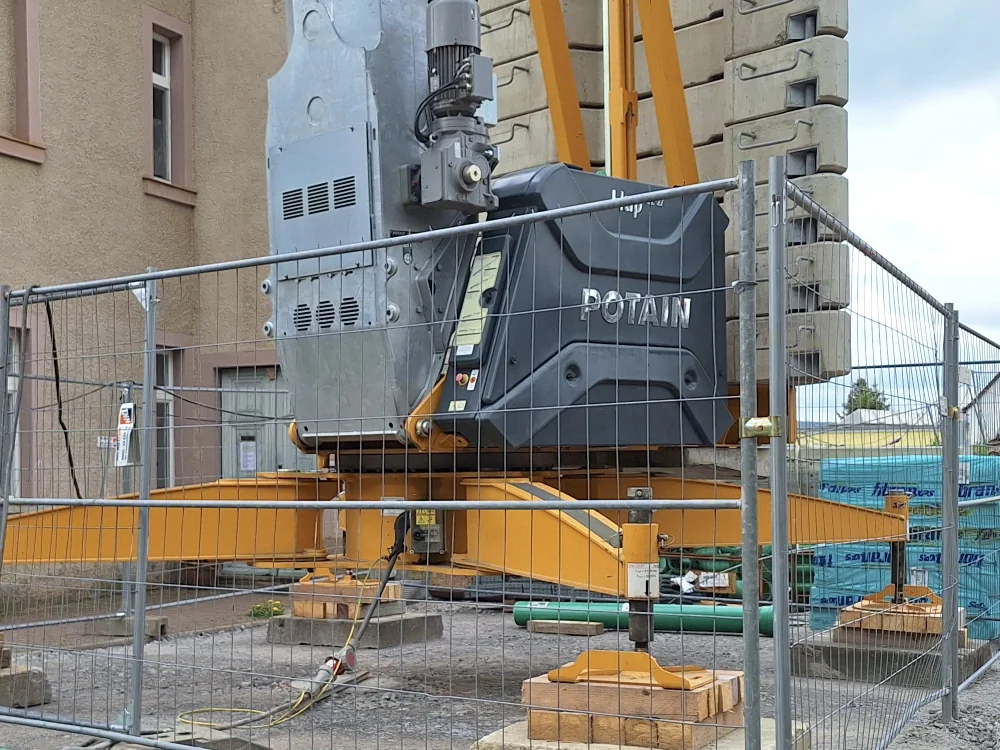 Baukran mit Ballastgewichten und Antriebseinheit auf Baustelle, umgeben von Bauzaun und Materiallager für Hochbauprojekt im Außenbereich.
