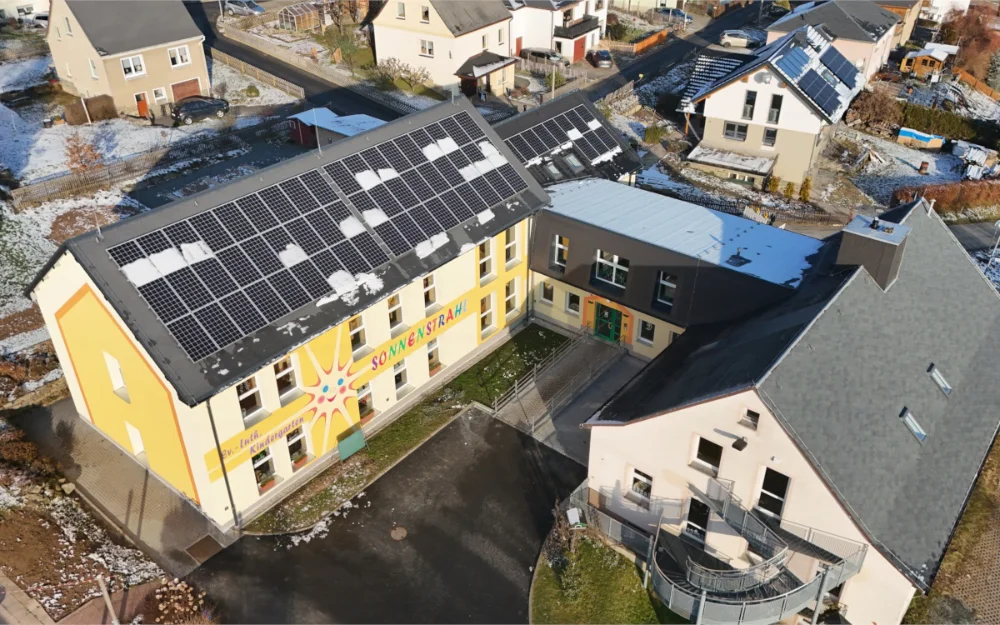 Drohnenaufnahme einer modernisierten Kindertagesstätte mit Photovoltaikanlage auf dem Dach, barrierefreiem Eingangsbereich und Außenanlagen in Wohngebiet.