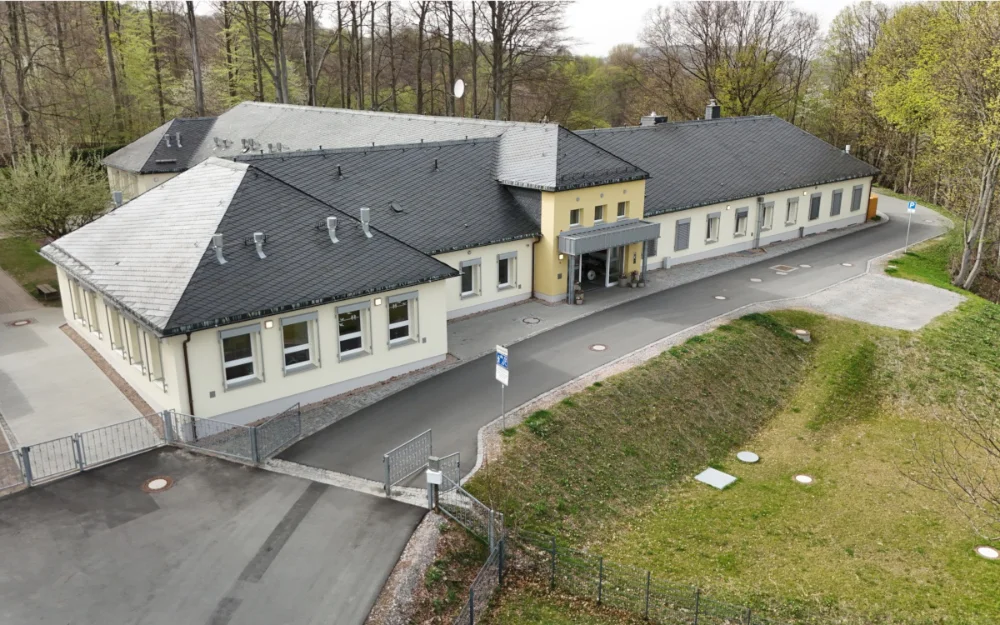 Drohnenaufnahme eines modernisierten Schulgebäudes mit neu gestalteter Zufahrt, Außenanlagen und barrierefreiem Eingangsbereich in naturnaher Lage.