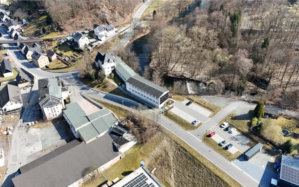 Drohnenaufnahme eines gemischt genutzten Gebiets mit Bestandsgebäuden, Parkplatzflächen und Infrastruktur entlang einer Straße nahe Flusslauf und bewaldeter Umgebung.