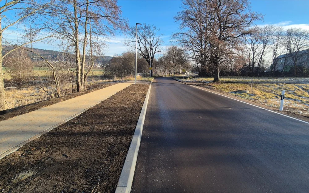 Neu ausgebaute Straße mit Asphaltfahrbahn, Bordsteinen und gepflastertem Gehweg, begleitet von Straßenbeleuchtung und Grünflächen im ländlichen Raum.